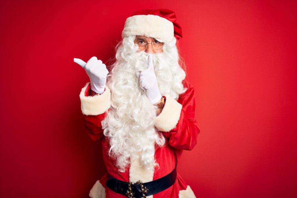 Middle age handsome man wearing Santa costume standing over isolated red background asking to be quiet with finger on lips pointing with hand to the side. Silence and secret concept. Smart Camels | Wij organiseren jouw Bedrijfsjubileum!